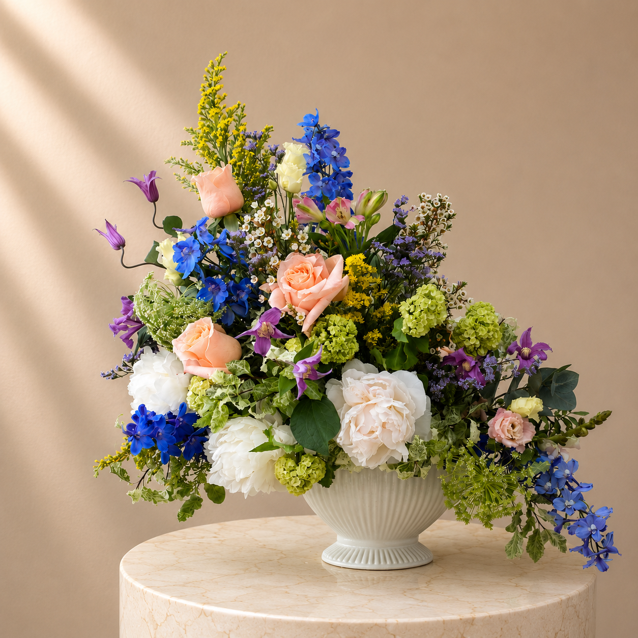 Sculptural seasonal arrangement in a white bowl with blue delphiniums, peach roses and soft green hydrangea
